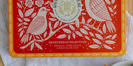A red and orange Shortbread Selection tin by Shortbread House is shown on a wooden table with flour, a spoon, and cookies on a plate. The tin features white floral and bird illustrations with the brand logo in the center.