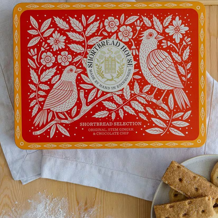 A red and orange Shortbread Selection tin by Shortbread House is shown on a wooden table with flour, a spoon, and cookies on a plate. The tin features white floral and bird illustrations with the brand logo in the center.