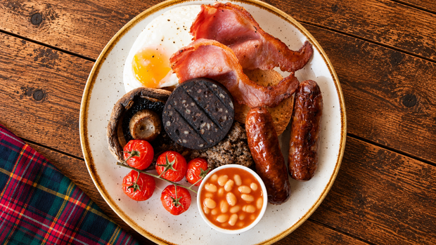 A full English breakfast served on a speckled beige plate with a gold rim, set on a rustic wooden table. The meal includes bacon, sausage, sausage, grilled black pudding, mushrooms, baked beans, and cherry tomatoes, with a small cup of baked beans in front.