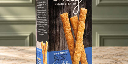A box of Stag Bakers Strathbann Blue Gourmet Cheese Straws, featuring a dark wood-textured black box with blue and white text and images of golden-brown cheese straws, set on a granite countertop with a blurred kitchen background.