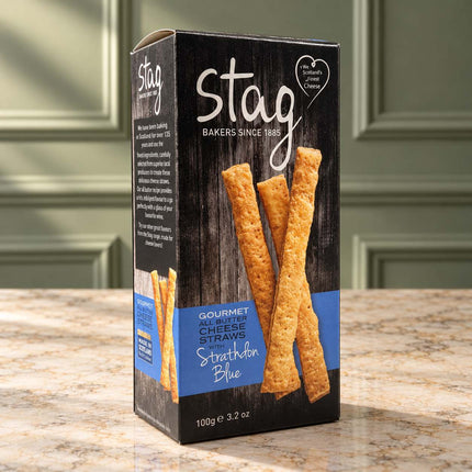 A box of Stag Bakers Strathbann Blue Gourmet Cheese Straws, featuring a dark wood-textured black box with blue and white text and images of golden-brown cheese straws, set on a granite countertop with a blurred kitchen background.