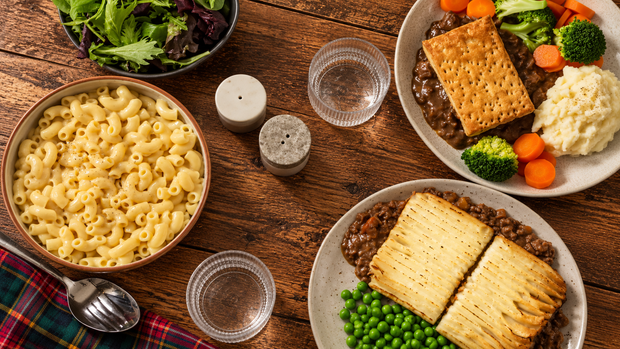 A rustic wooden table styled with a hearty meal, featuring a bowl of macaroni pasta, two plates of shepherd's pie with crackers, broccoli, carrots, mashed potatoes, peas, and a side salad.